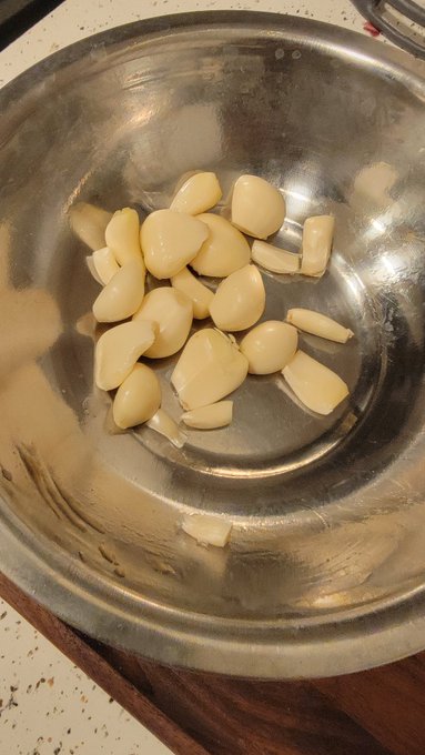 A stainless steel bowl contains peeled garlic cloves resting on a wooden cutting board.