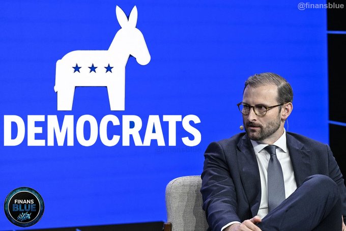 A bearded man in a dark suit and tie sits on stage at a 'DEMOCRATS' event, with the party's donkey logo displayed on the blue screen behind him and 'FINANS BLUE' watermark visible.