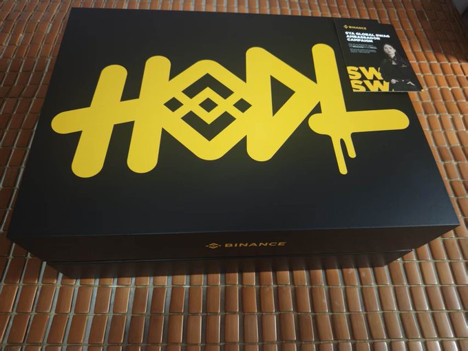 A black and yellow Binance-branded box displaying the graffiti-style text "HODL" sits on a tiled surface, accompanied by a promotional card for the "BTS Global Dubai Activation Campaign" featuring SWAY. The image ties into the celebratory post marking Binance's 9th anniversary, highlighting BNB's growth from a $0.15 ICO price to $570 and the exchange's pioneering journey through crypto milestones.