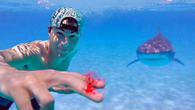 An underwater selfie of a smiling man wearing a patterned swim cap, with a shark visible in the blue water behind him.
