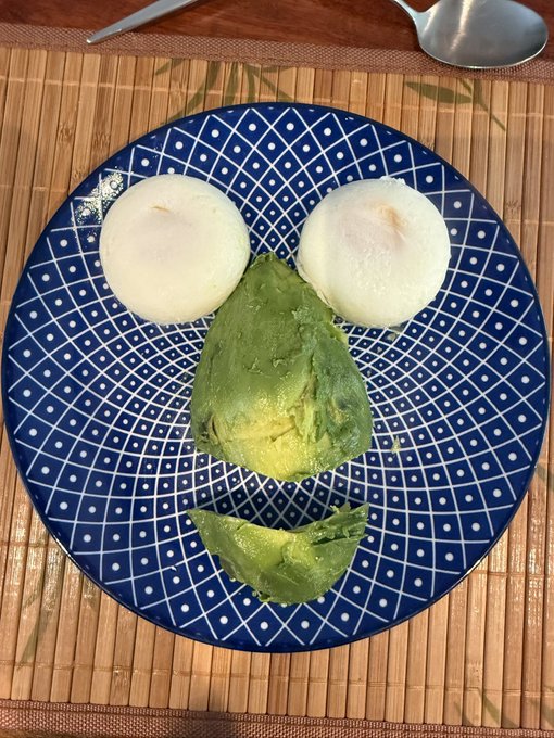 A creative food arrangement on a blue and white patterned plate featuring two white round items as eyes, a green avocado piece as a nose, and another piece as a smiling mouth, creating a face design.