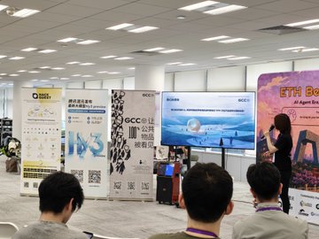 The image shows a group of people gathered around a large screen, possibly watching a presentation or learning about a specific topic. There are at least five individuals visible in the scene, with one person standing near the center-left side of the frame and others scattered throughout the room. The main focus is on the screen, which appears to be displaying information relevant to the event taking place.