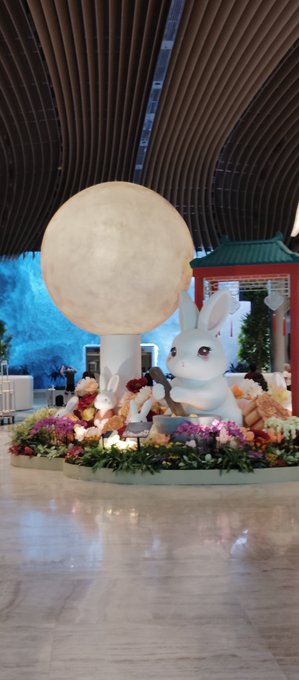 A large illuminated sphere sits beside an oversized white rabbit figurine holding a wooden mallet, surrounded by a colorful flower display and a red traditional-style pavilion in an indoor space.