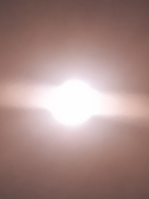 An overexposed, blurry photo of a bright light source against a pale, pinkish-beige background.