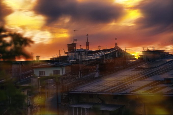 A dramatic sunset with golden and dark clouds illuminates a cityscape of rooftops, antennas, and a domed church.