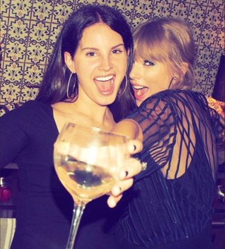 Two women—one with dark hair and one with blonde hair—cheerfully clink wine glasses at an upscale venue with ornate patterned wallpaper. The image appears to celebrate the rumored friendship between the two pop stars, hinting at the lively celebration that might unfold at Swift's reported wedding.