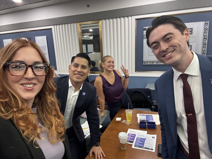 A selfie taken indoors showing four people smiling at the camera, with a woman wearing tortoiseshell glasses in the foreground left and a man in a blue suit on the right.