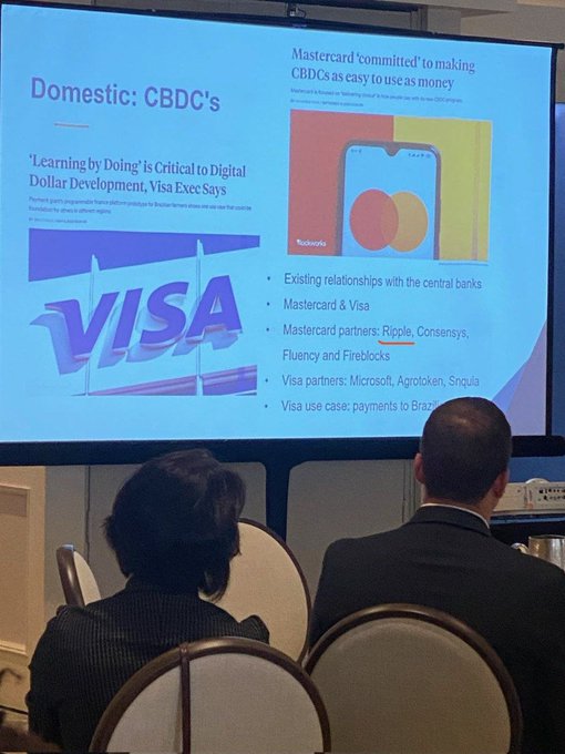 The image shows a presentation screen with two people sitting behind a table, watching a presentation about Mastercard's new crypto debit card. The card allows users to spend from self-custodial wallets at 100 million merchants across Europe. The focus of the presentation is on the Mastercard's new initiative and how it can be used for making payments using cryptocurrency.
