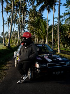 A person in a black hoodie and red cap leaning against a black rally car with sponsor decals, posed on a road lined with tall palm trees.