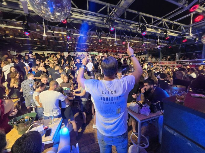 A crowd gathers at a lively nightclub or event venue where a person in the foreground, wearing a "Czech Liquidity Provider" shirt, raises their arms with a glowing blue light stick, while the "LFG @SolanaCZE" caption suggests this is a Solana community gathering in the Czech Republic.