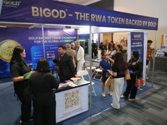 The image shows a promotional booth for BIGOD, described as "THE RWA TOKEN BACKED BY GOLD," at what appears to be a crypto or financial event. Staff members are engaging with attendees at the booth, which features gold token imagery and display materials, supporting the post's announcement of a 7.7 MEGA AIRDROP giveaway to celebrate the token's campaign.