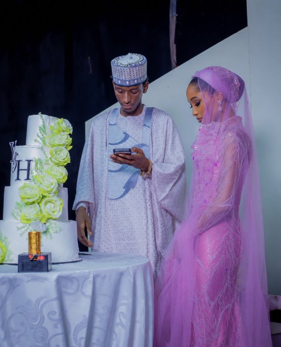 A groom in traditional attire checks his phone while standing beside his bride in a pink dress next to a multi-tiered wedding cake decorated with green roses and the letter "H," humorously illustrating the caption "Let me buy this $SUNUSI dip first."