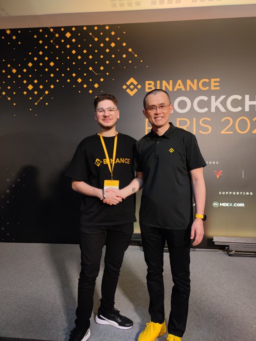 Two men pose at a Binance Blockchain Paris 2025 event, with one wearing a Binance t-shirt and the other in a black polo.