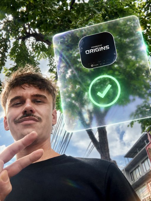A young man with a mustache poses outdoors under trees while holding up a holographic NFT card labeled "TOKENIZED ORIGINS" displaying a checkmark.