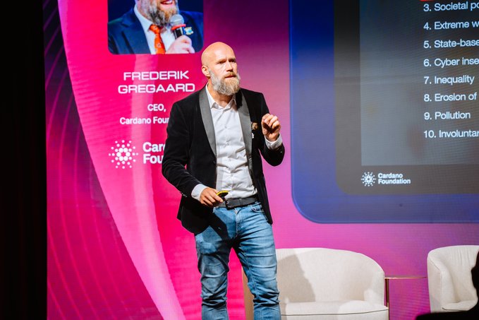 Frederik Gregaard, CEO of Cardano Foundation, presents on stage with a backdrop listing global challenges including societal, extreme weather, cyber insecurity, inequality, and pollution.
