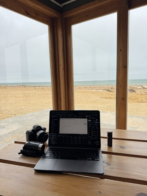 A laptop displays trading charts and market data while sitting on a wooden surface next to a DSLR camera, with an overcast ocean view in the background through large windows—capturing a coastal remote workspace setup, fitting for a "GM $LARRY" good morning greeting.