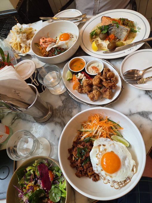 An overhead view of a marble table laden with various restaurant dishes, including bowls topped with fried eggs, fried chicken pieces, salads, and a plate with fish.