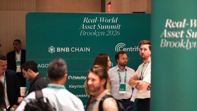 Attendees network at the Real-World Asset Summit in Brooklyn 2026, with the event's teal backdrop prominently featuring sponsor logos including BNB Chain, Centrifuge, and Fidelity.