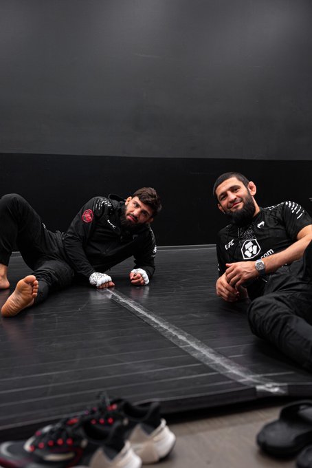 Two bearded men wearing black Crypto.com-branded athletic gear sit on a black mat with a strip of tape between them, appearing relaxed yet intense as they pose for the camera, while several pairs of athletic shoes are scattered in the foreground—fitting the accompanying post's promotional tie-in to Crypto.com, UFC 331, and the PPlusDeportes broadcast.