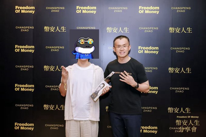 Two men pose in front of a backdrop displaying 'Freedom Of Money' and 'CHANGPENG ZHAO' in both English and Chinese ('幣安人生'); one man's face is obscured by a cartoon sticker.