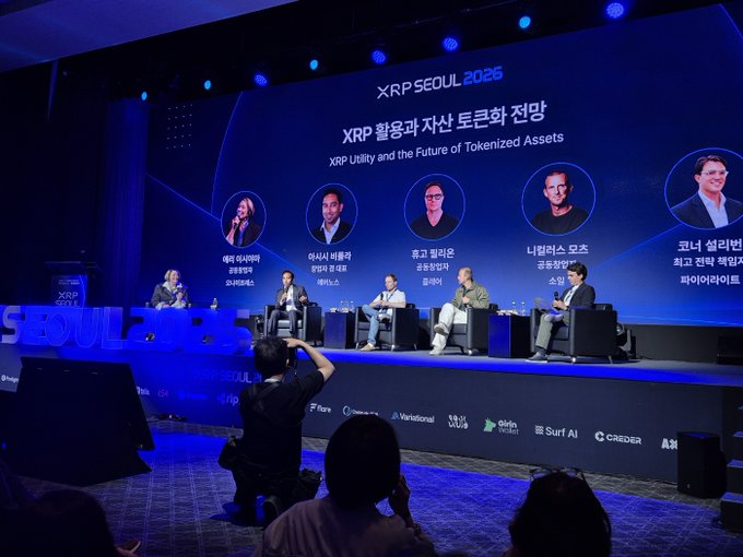 A panel discussion at XRP Seoul 2026 titled "XRP Utility and the Future of Tokenized Assets" features speakers on stage discussing the expansion of XRP into DeFi and tokenized finance, with a photographer capturing the event from the floor. The image directly illustrates the post's theme of institutional-grade infrastructure development on the XRP Ledger, particularly highlighting Korea's prominent role in the XRP ecosystem.