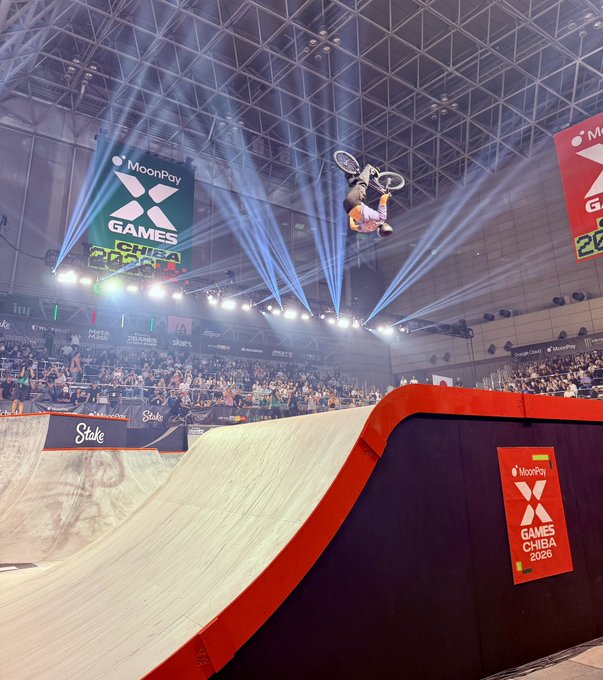 A BMX rider executes an inverted aerial trick mid-flip above a large vert ramp at the MoonPay X Games Chiba 2026 event, with dramatic spotlights and "Stake" sponsor banners visible below.

Caption: "MOON THAT" — capturing the gravity-defying flip the rider is throwing above the MoonPay-branded vert ramp at X Games Chiba 2026, with the massive MoonPay Games Chiba banner looming overhead as if the rider is flipping toward the moon itself.