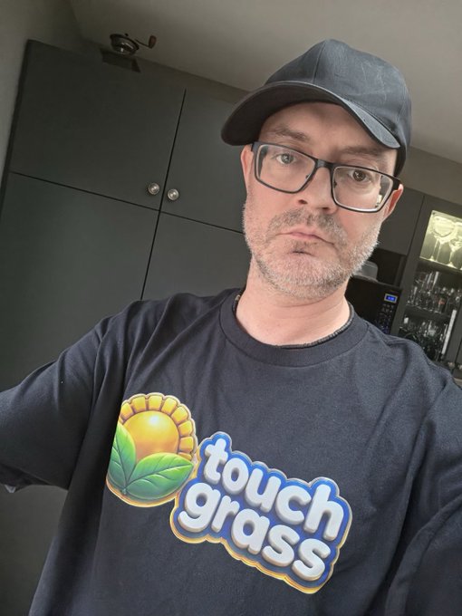 A man wearing glasses, a black cap, and a black "Touch Grass" t-shirt poses for a selfie, matching the post's description of someone repping the brand at Blockchain Futurist.