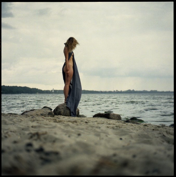 A photograph of a person with curly blonde hair standing on a rock at the beach, draped in a blue cloth, with a cloudy sky and water in the background.