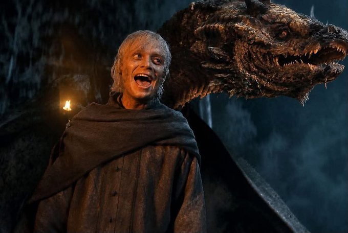 A still from a television show showing a man with pale skin and blonde hair in a brown cloak smiling, standing in front of a large, menacing dragon's head in a dark, cave-like setting.