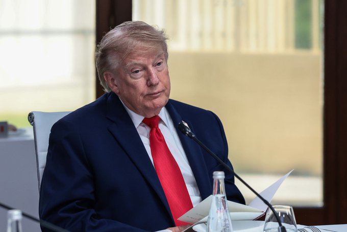Donald Trump is seated at a table wearing a dark suit and red tie, with a microphone positioned in front of him.