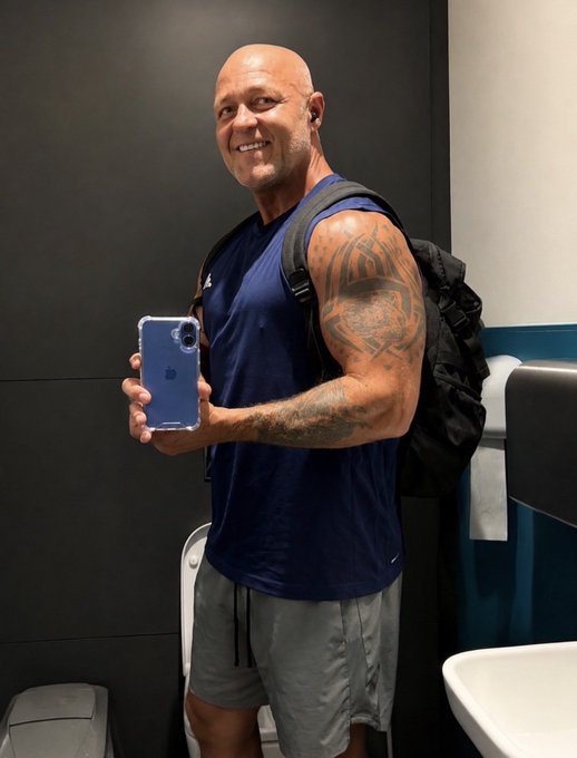 A smiling bald man in a navy blue tank top and gray shorts stands in a restroom, showing off his phone while wearing a backpack and flexing his tattooed arm. The image reflects the post's "Gym smashed" caption, capturing a triumphant post-workout moment with his fitness buddy Treb.