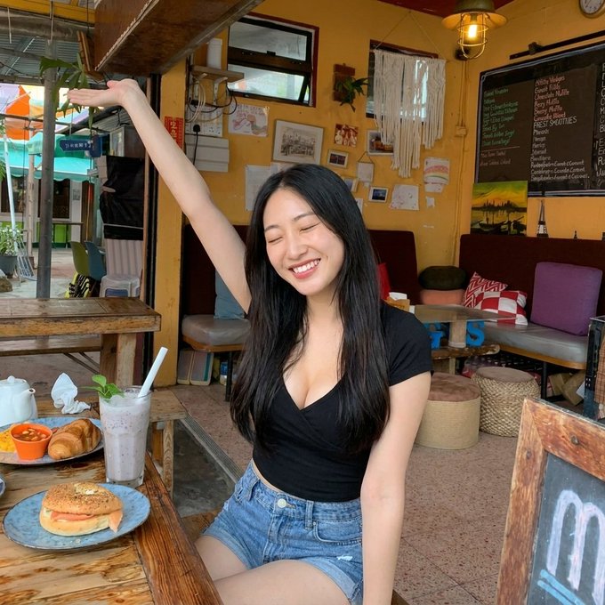 A smiling young woman with long dark hair sits at a wooden table in a cafe, with a burger and a drink in front of her.