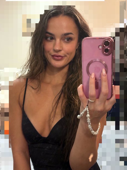A mirror selfie of a young woman with long brown hair wearing a black top, holding a pink iPhone with a pearl phone charm, with pixelated areas obscuring parts of the background.
