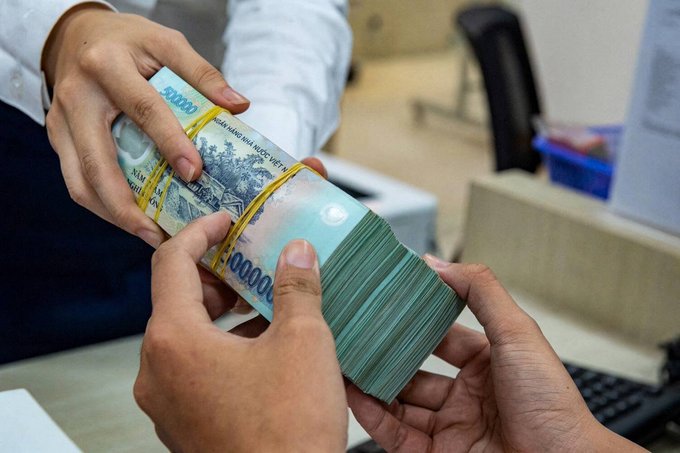 Hands hold a thick stack of Vietnamese dong banknotes bundled together with yellow rubber bands in an office setting.