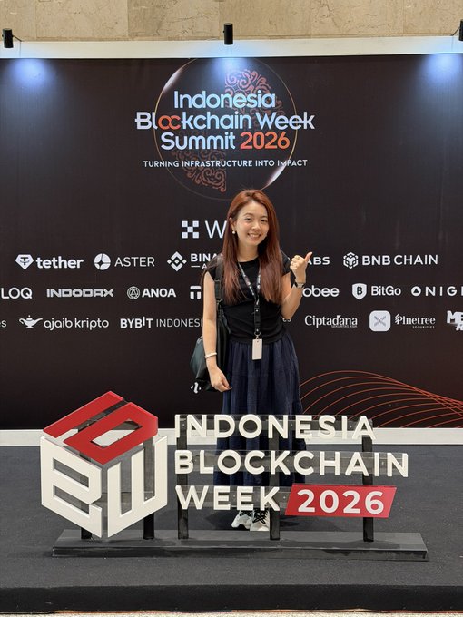 A woman posing and giving a thumbs-up in front of an 'Indonesia Blockchain Week Summit 2026' backdrop with the slogan 'TURNING INFRASTRUCTURE INTO IMPACT' and various sponsor logos.