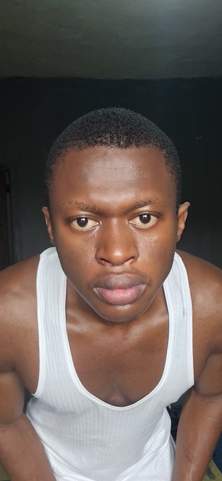 A close-up selfie of a young man wearing a white tank top against a dark background.
