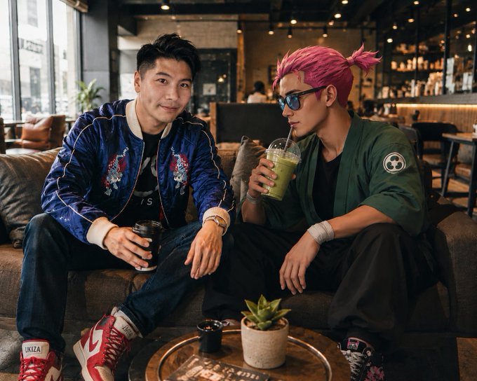 Two young men sitting on a leather couch in a cafe/lounge; one wears a blue embroidered bomber jacket with red and white sneakers, while the other has pink hair tied back, blue sunglasses, a green jacket, and drinks a green smoothie. A small potted succulent sits on the coffee table in front of them.