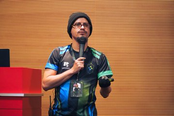 A man wearing a beanie, glasses, and a blue and black gaming jersey speaking into a microphone on a stage.