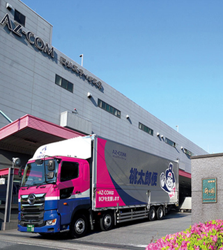 A pink and purple AZ-COM Maruwa HD delivery truck is parked outside the company's headquarters, displaying Japanese text reading "橋本運輸" (Hashimoto Transport), visually representing the logistics firm that is partnering to adopt JPYC stablecoin payments for its network of approximately 2,300 partner companies.