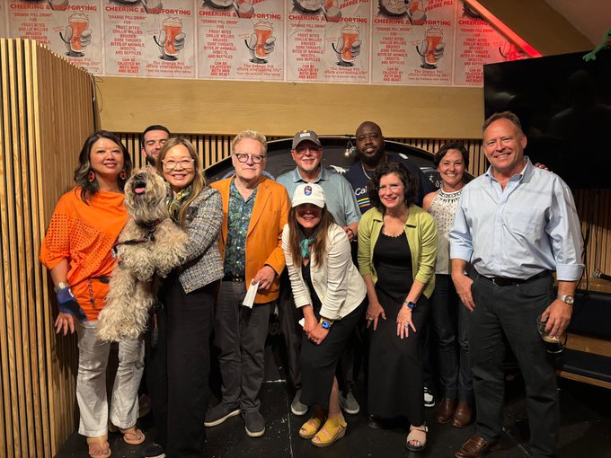The image features a group of people posing for a photo, with some standing close together while others are more spread out. A man wearing an orange shirt can be seen holding a dog, which adds a playful element to the scene. The group appears to be gathered in front of a wall adorned with posters and signs, possibly indicating that they have just attended or participated in an event like the Pitch at Pubkey in NYC.