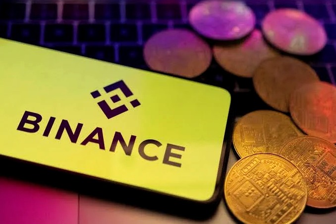 A smartphone displays the yellow Binance app logo alongside several gold-colored physical Bitcoin tokens resting on a laptop keyboard.