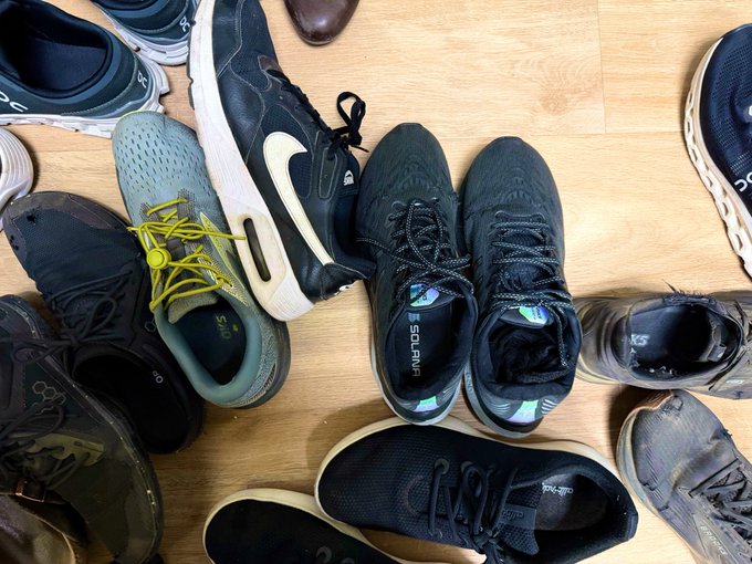The image shows a collection of various shoes, including sneakers and dress shoes, scattered across a wooden floor. The shoes are arranged in different positions, with some standing upright while others lie flat or appear to be lying down. There are at least 13 pairs of shoes visible in the scene, showcasing an assortment of styles and colors. The image does not provide any information about the location or context of these shoes, but it appears that they might have been left out on a wooden floor for some time.