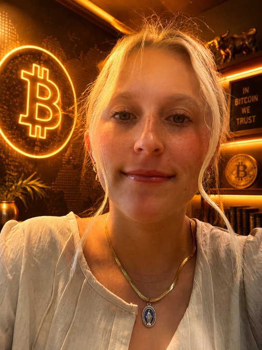A blonde woman takes a selfie in front of glowing neon Bitcoin signage and a sign reading "IN BITCOIN WE TRUST," embodying the post's message about Bitcoiners' dedication to understanding and escaping the traditional financial system. The warm, orange-lit Bitcoin-themed backdrop visually reinforces the post's theme of trust in Bitcoin over conventional currency.