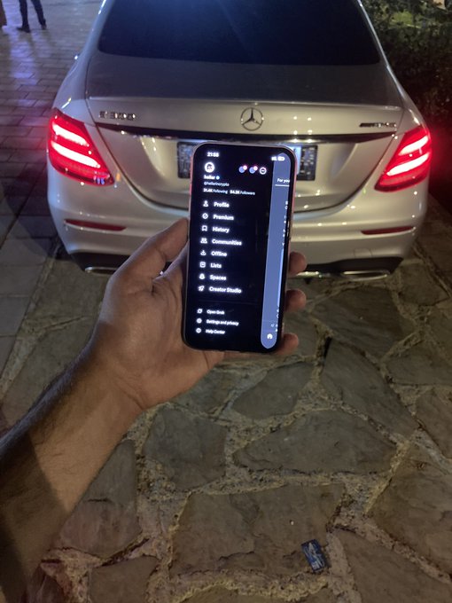 A hand holds up a smartphone displaying a streaming app's content categories (Profile, Premium, History, Communities, etc.) in front of a parked silver Mercedes-Benz at night.