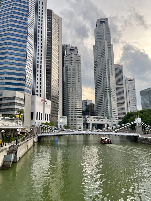 A view of the Singapore River with modern skyscrapers, a pedestrian bridge, and a small boat on the water.