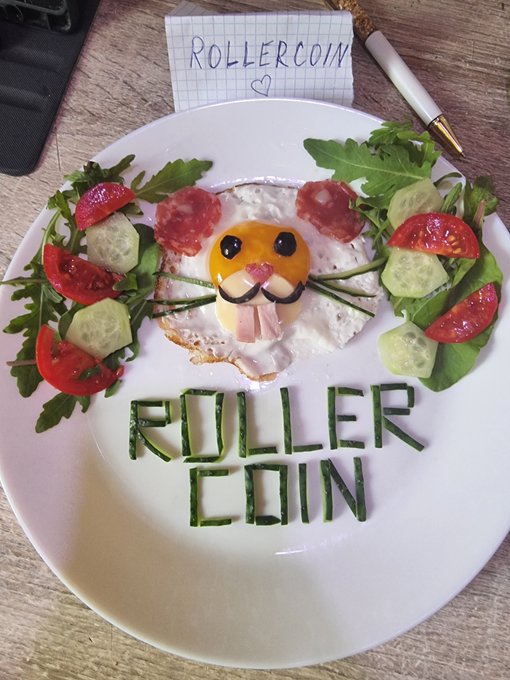 A creative food art plate spelling "ROLLERCOIN" with green vegetables, featuring a smiley face made from egg, cucumber, and salami, along with fresh vegetables, garnished with a "ROLLERCOIN" note card.