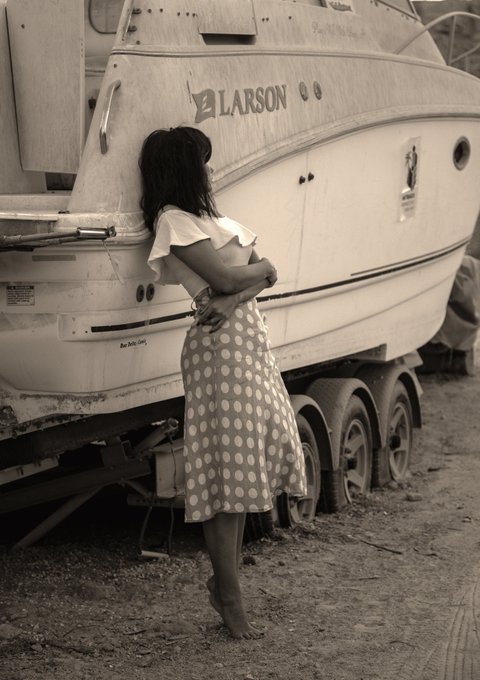 A sepia-toned photograph shows a woman in a polka-dot dress leaning against a white 'LARSON' boat on a trailer, with her face obscured.