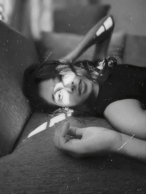 A black-and-white artistic portrait photograph of a woman lying on a couch with dramatic lighting streaming across her face, her hand extended toward the camera, with visible dust particles in the air.