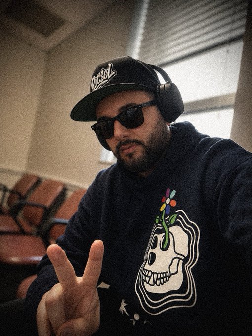 A selfie-style photo of a man wearing a black 'Ursol' cap, sunglasses, headphones, and a black hoodie with a flower-skull design, flashing a peace sign.