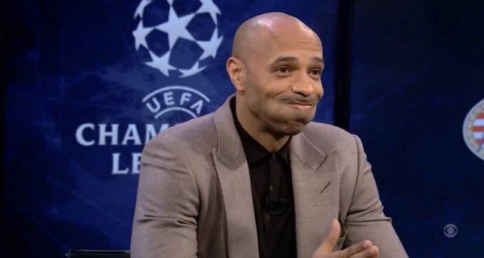 A bald man in a gray suit and black shirt sits in front of a UEFA Champions League backdrop, appearing to be on a sports broadcast panel.